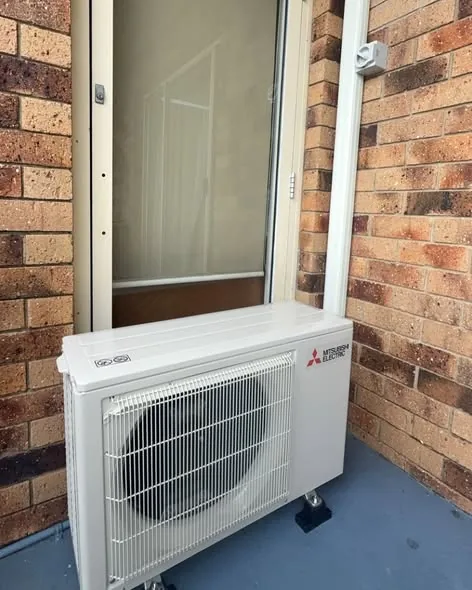 Mitsubishi split-system outdoor unit standing level on mounting feet beside a brick wall
